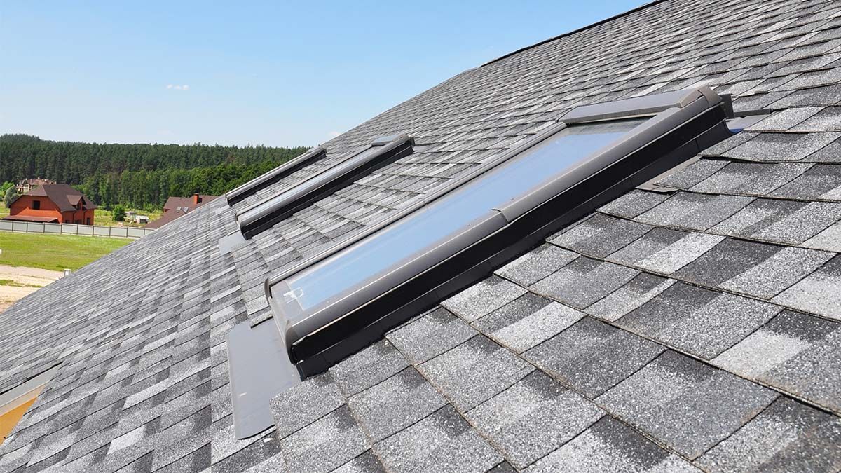 Three rectangular skylights on a gray shingle roof under a blue sky, overlooking a landscape.