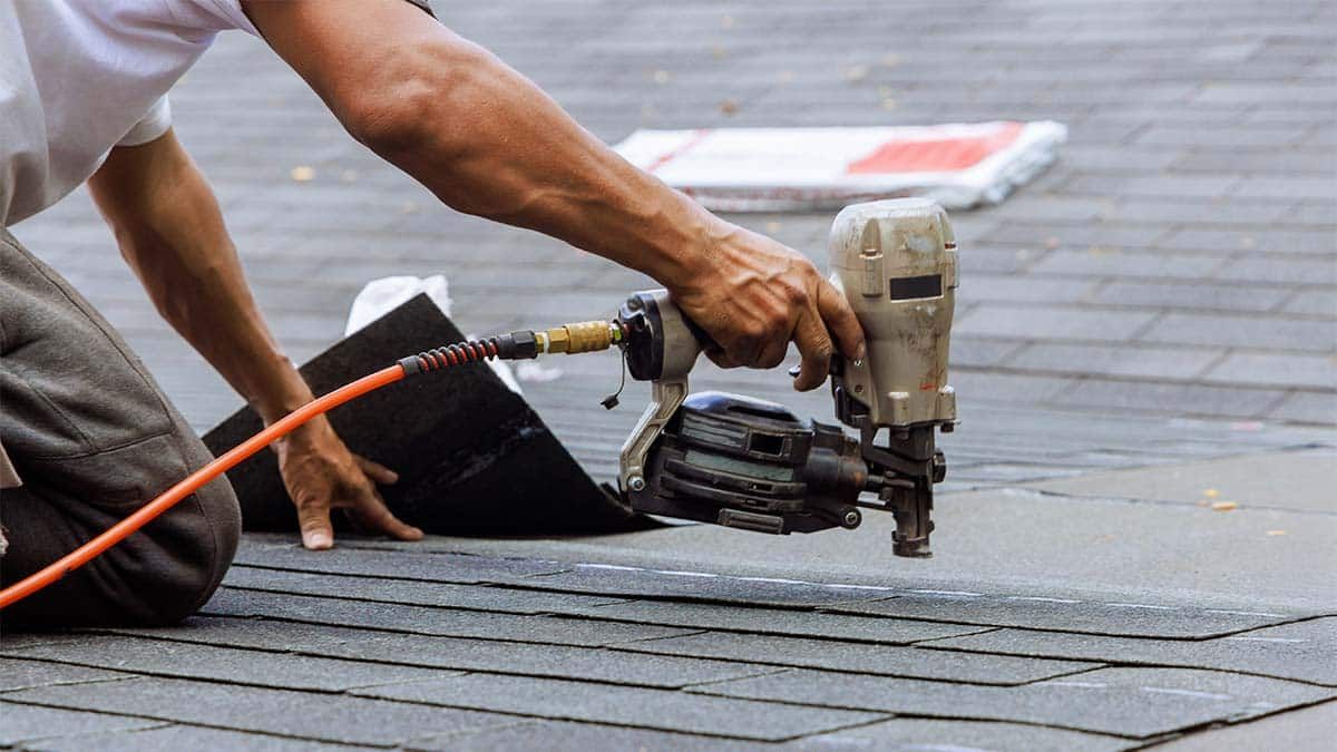 A person kneeling on a roof, using a nail gun to install roofing shingles.