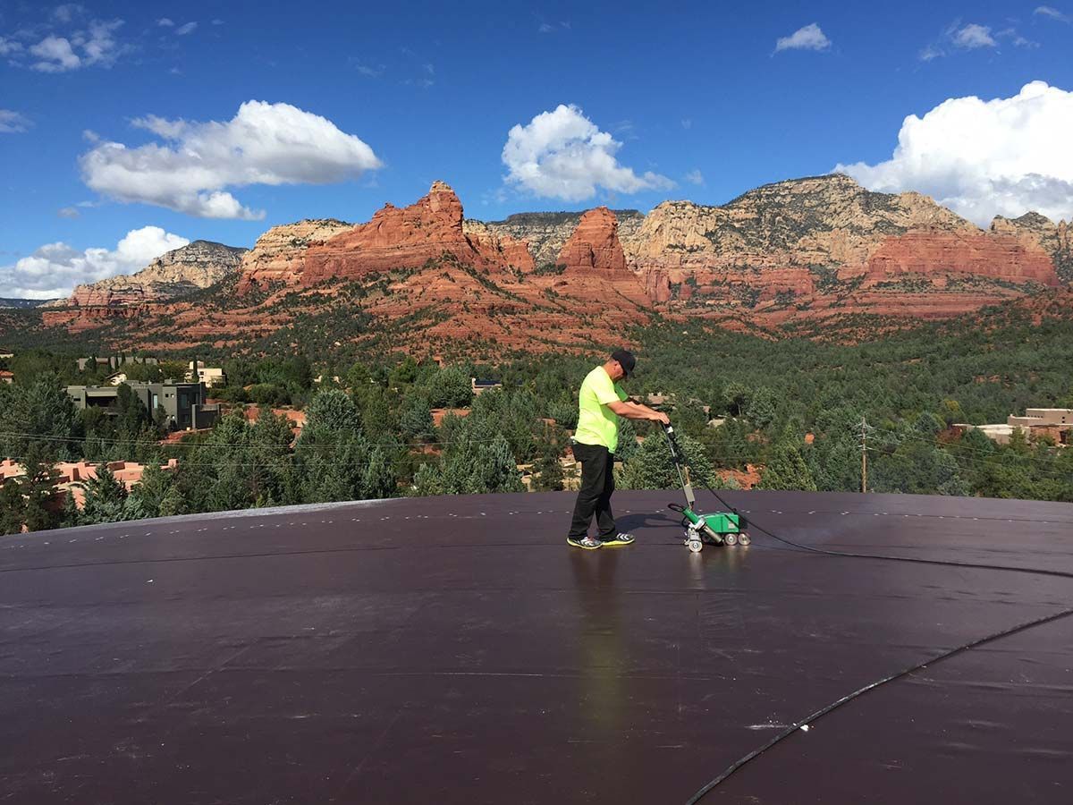 Man cleaning a dark roof with red rock mountains in the background under a blue sky with clouds.