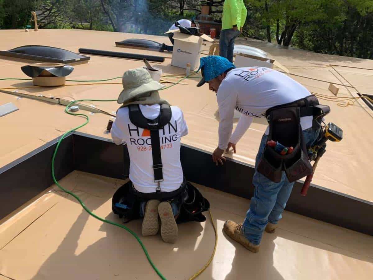 Two roofers installing a dark edge on a flat roof. One kneels and the other bends, both wearing safety gear.