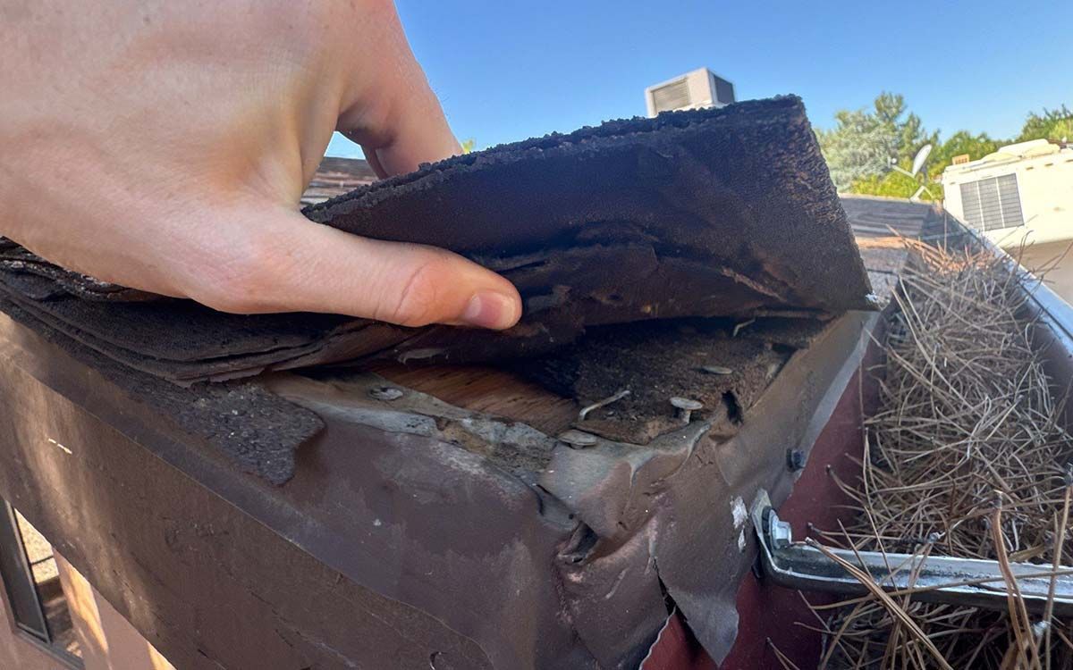 Hand lifting a damaged asphalt shingle on a brown gutter, revealing debris and potential roof damage against a blue sky.
