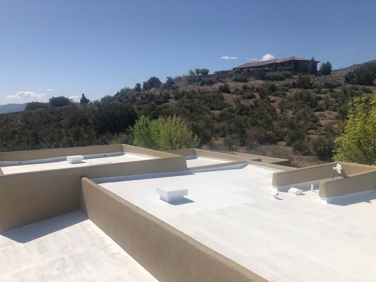 Flat roof with white coating, beige parapets. Distant hillside with trees and building. Blue sky.