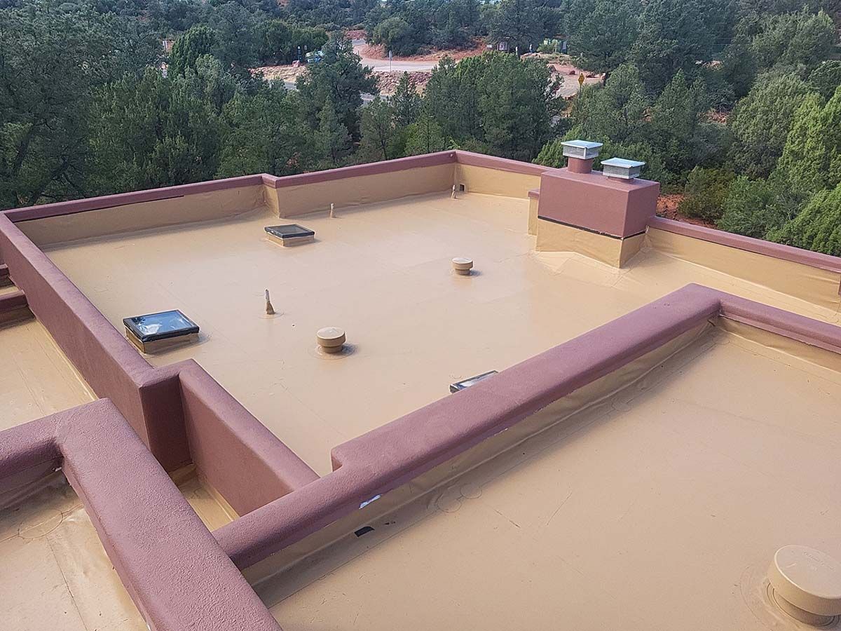 Flat rooftop with tan surface, brown trim, and several vents, surrounded by green trees.