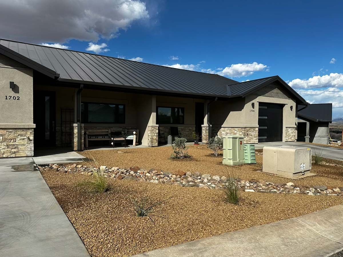Single-story beige building with black roof and garage. Landscaped front yard with rocks and drought-tolerant plants. 