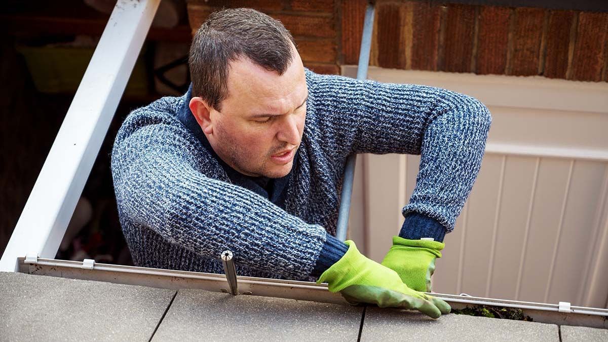 Man installing gutter guards on a house roof, wearing gloves and a blue sweater.