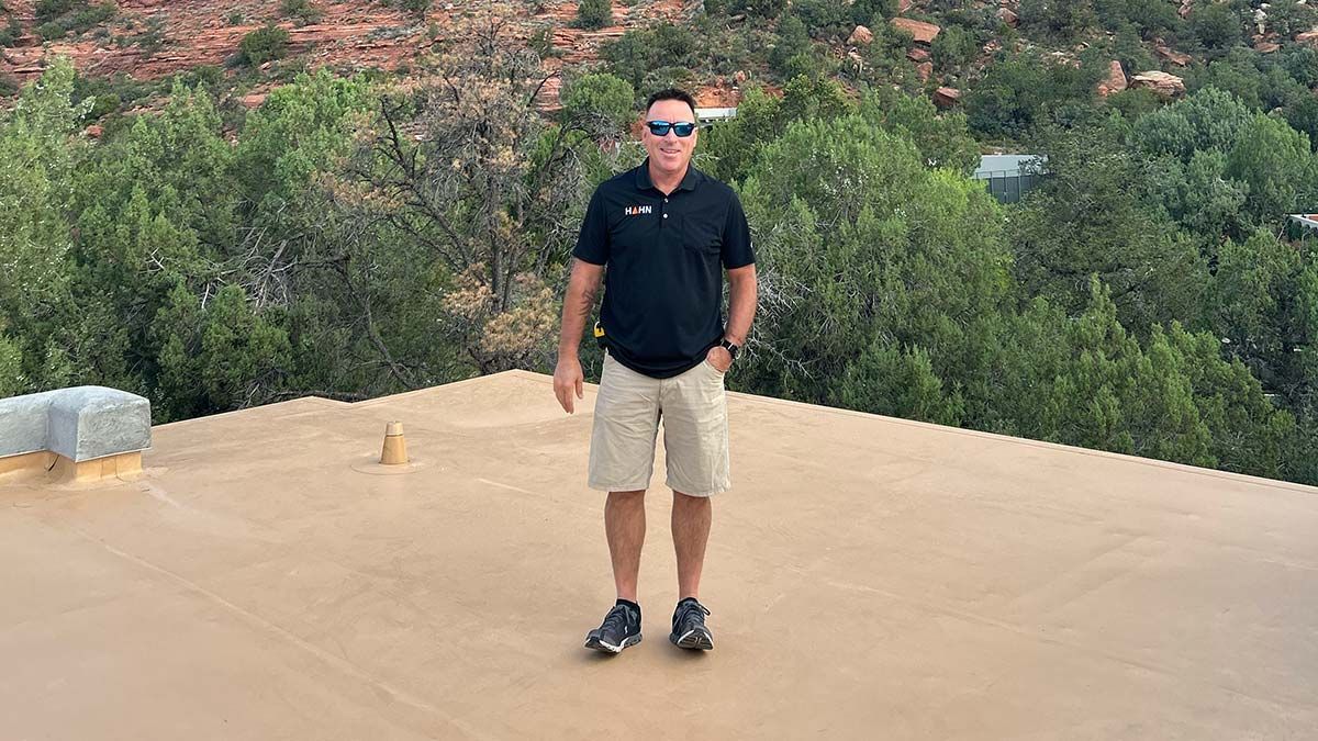 Man in black shirt and khaki shorts on a flat, brown roof with a green tree-filled background.