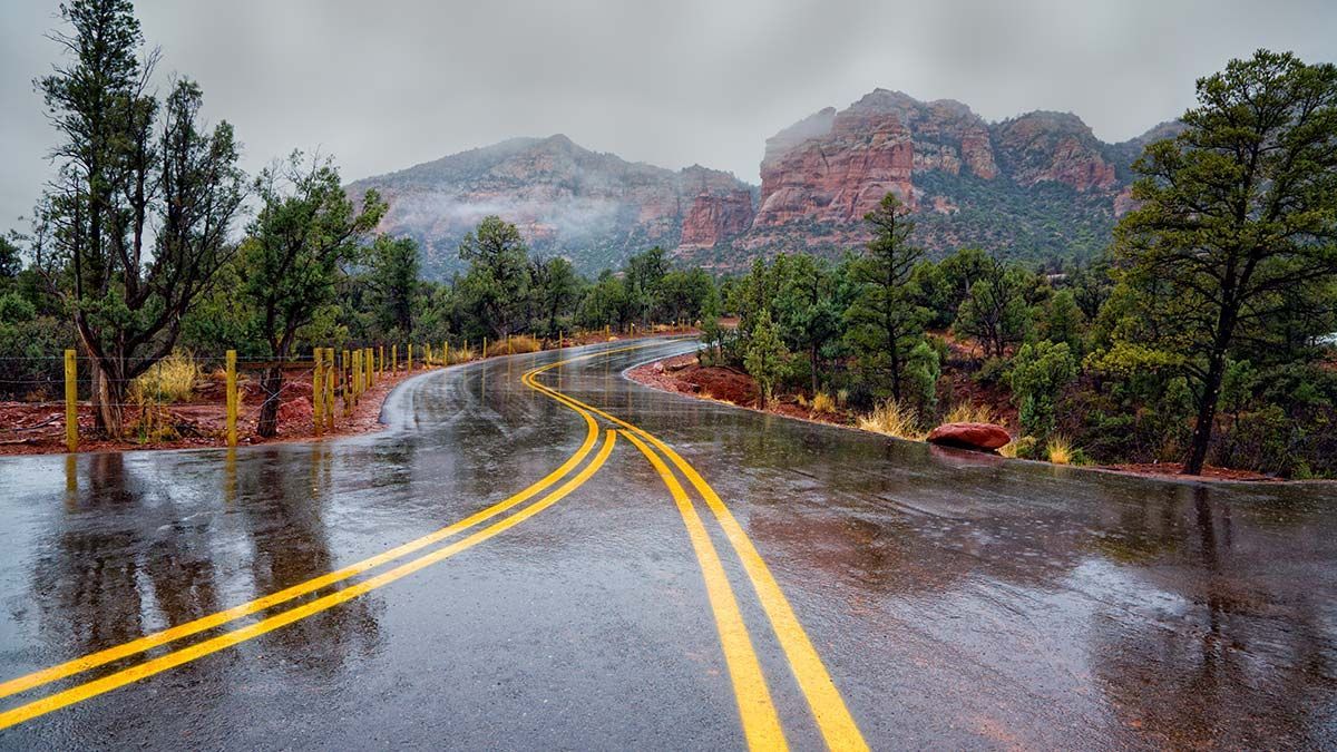 Wet road winding through trees toward red rock mountains in the rain.