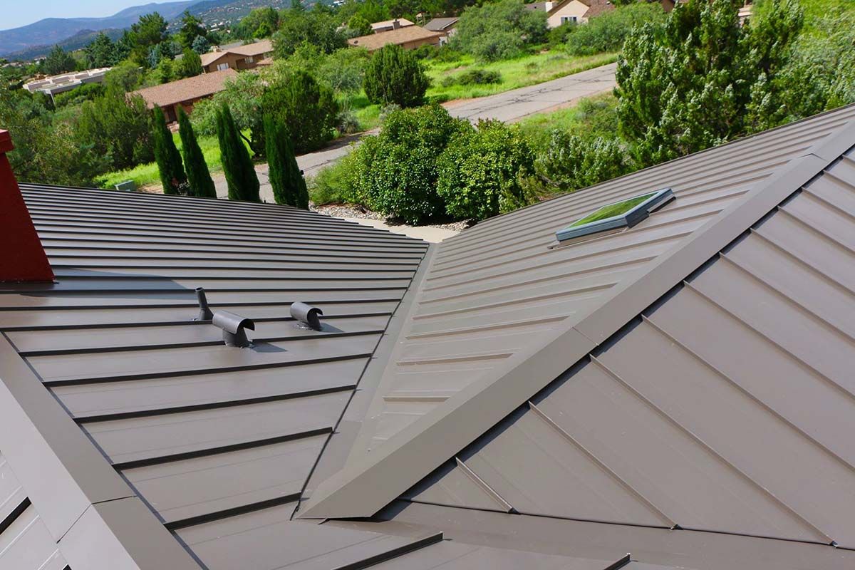 A brown metal roof with visible ridges, chimneys, and vents, set against a backdrop of greenery and houses.