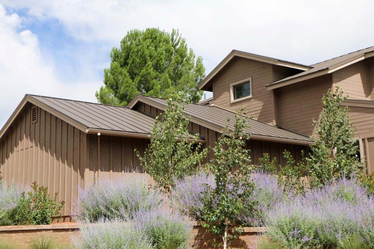 Brown house with metal roof, surrounded by purple flowers and trees, under a blue sky.