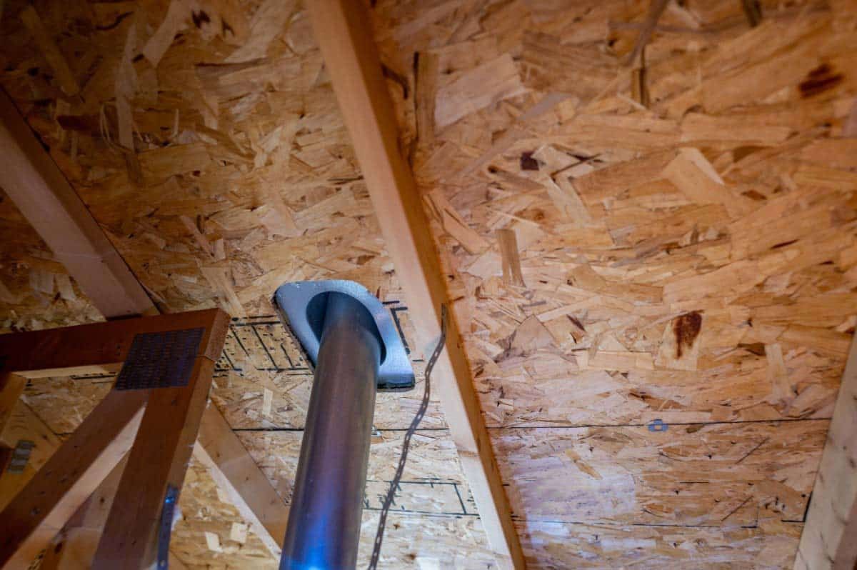 An upward-facing view inside an attic shows a metal pipe passing through wooden framing and oriented strand board sheathing.