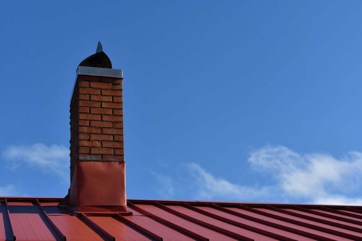 Brick chimney on a red metal roof against a bright blue sky with a few clouds.