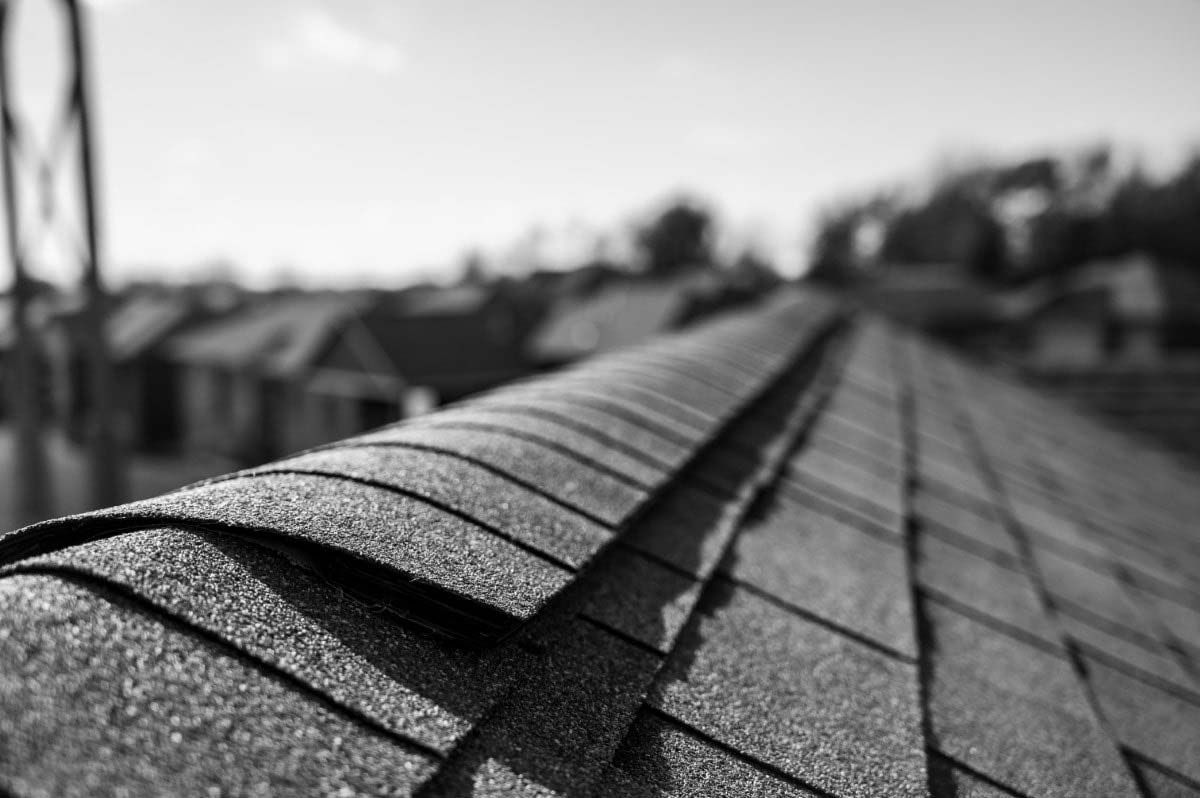 Close-up of a roof with shingles, leading to a blurred neighborhood backdrop in black and white.