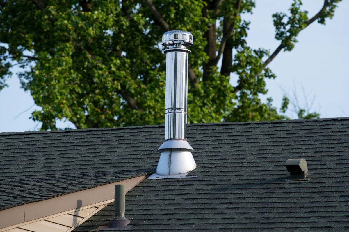 Metal chimney extending from a dark shingled roof against a backdrop of green trees.
