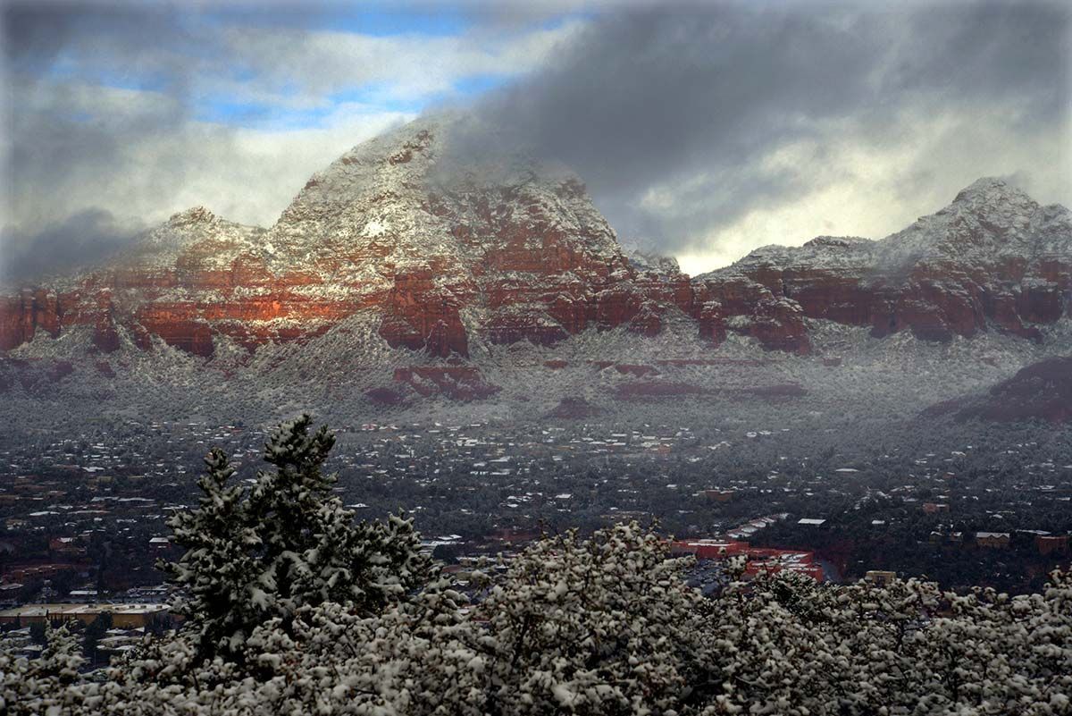Snowy red rock mountains with a hazy town in the valley, under cloudy skies.