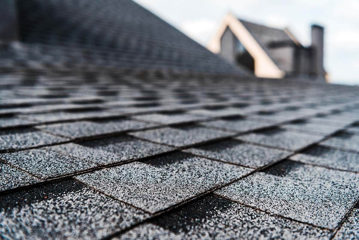 Close-up of asphalt shingles on a roof, leading to a house in the background.