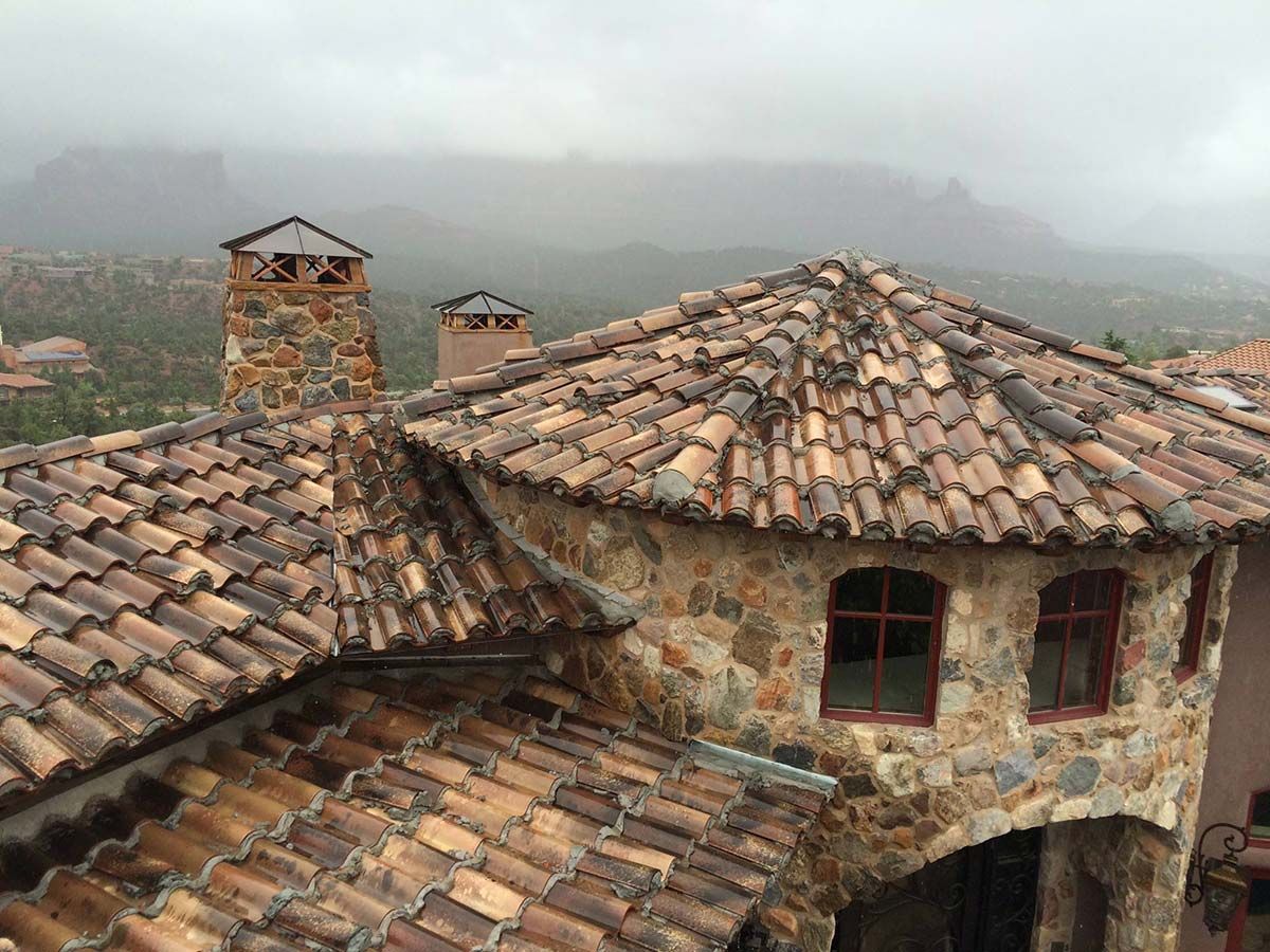 Stone building with clay tile roof and chimneys, mountains in the background under a cloudy sky.