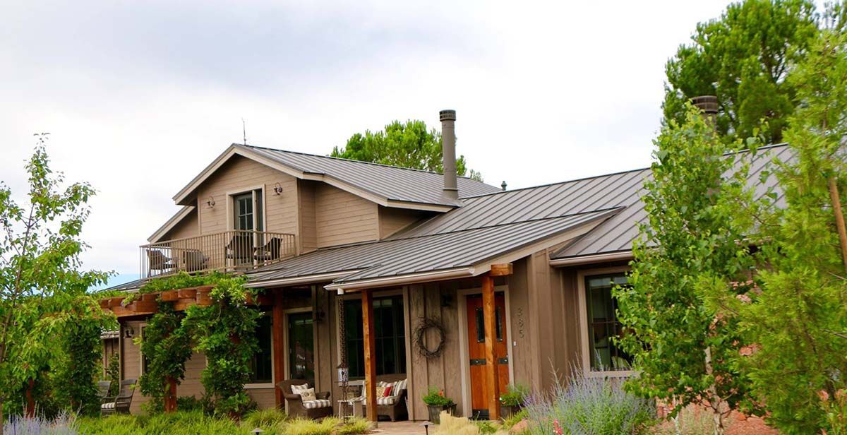 Tan adobe-style house with metal roof, surrounded by green trees and bushes, under a cloudy sky.