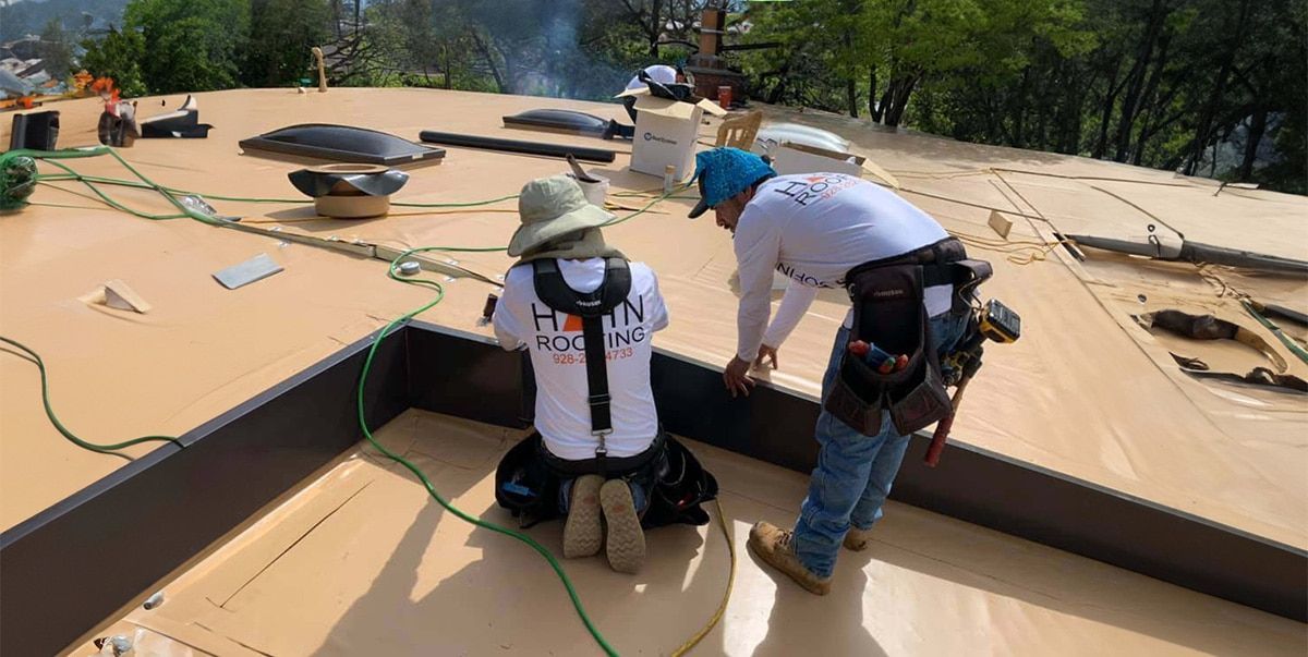 Two construction workers installing roofing on a flat roof. One kneels, the other stands.