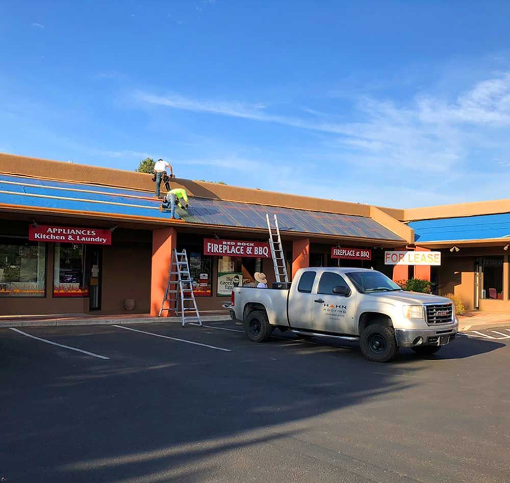 Workers installing solar panels on a building's roof with a pickup truck parked below. Clear blue sky.