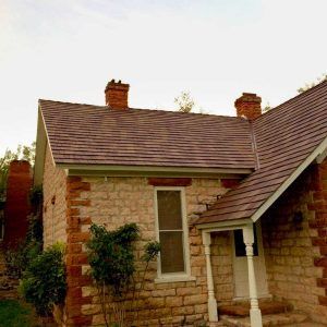 Stone house with red brick chimneys and roof. Door and window visible, with some greenery around.