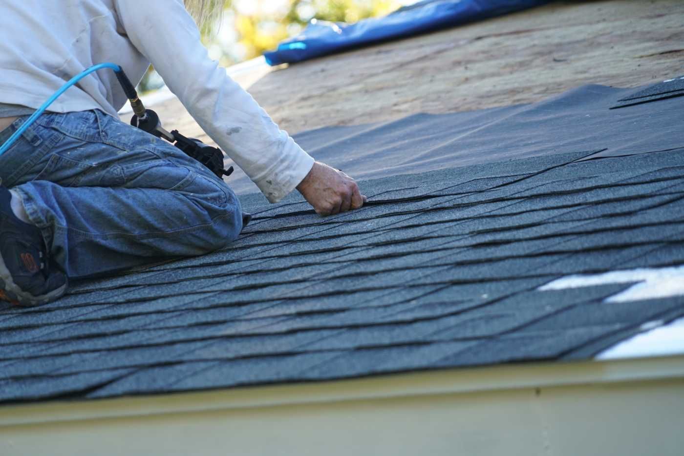 Roofer on a roof, installing dark shingles.  A blue tarp is visible in the background.