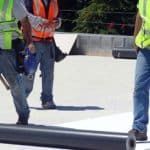 Construction workers in safety vests on a flat roof, working near black piping.