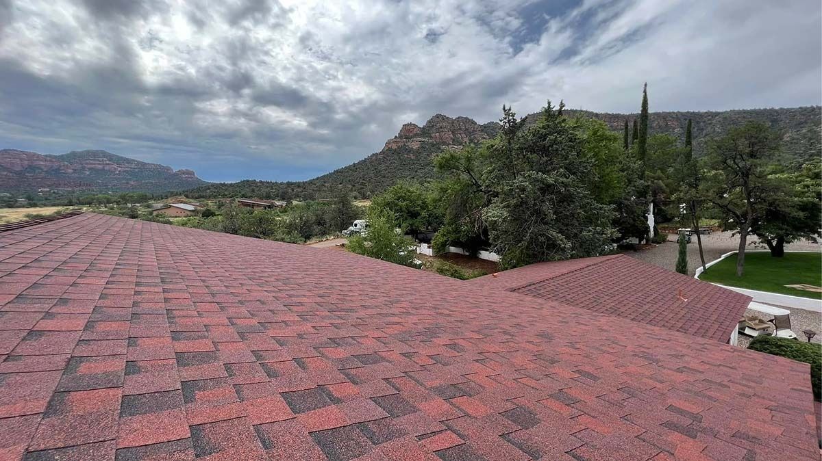 Red asphalt shingle roof with a scenic backdrop of trees, mountains, and a cloudy sky.