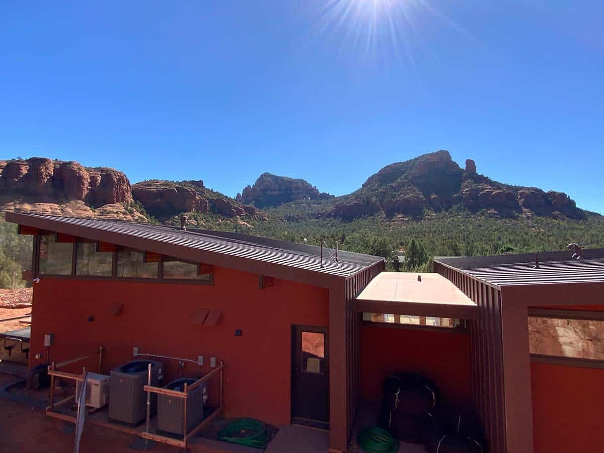 Red building with solar panels, facing red rock mountains under a sunny blue sky.