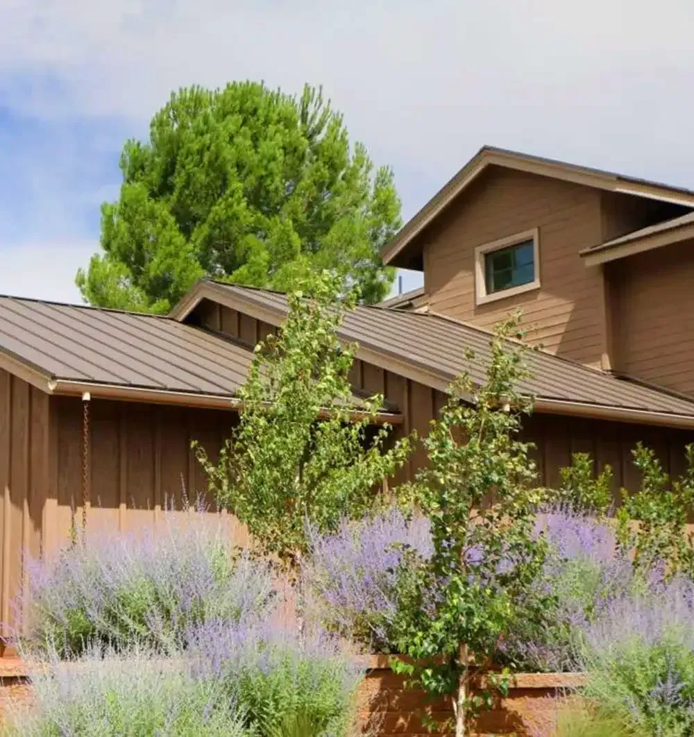 Brown house with metal roof, lavender bushes, and green trees against a blue sky.