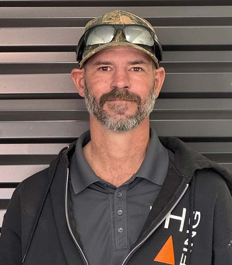 Man wearing a camouflage cap and glasses, gray polo shirt, and black jacket, in front of a gray wall.