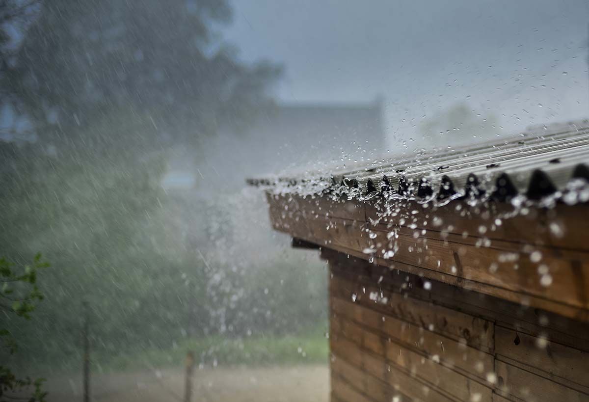 Rain falling from a wooden roof with water droplets visible. Green foliage in the blurry background.
