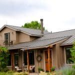 Tan-colored house with a dark gray roof and chimney, a small porch, and greenery in front.