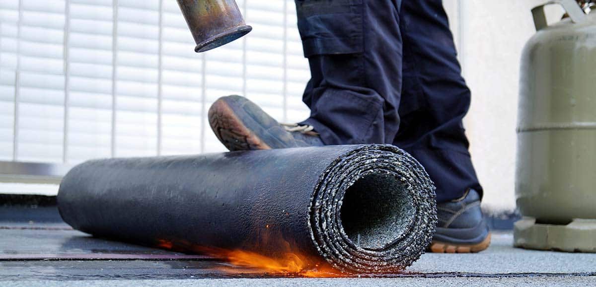 A person using a torch to melt a roll of roofing material on a rooftop.