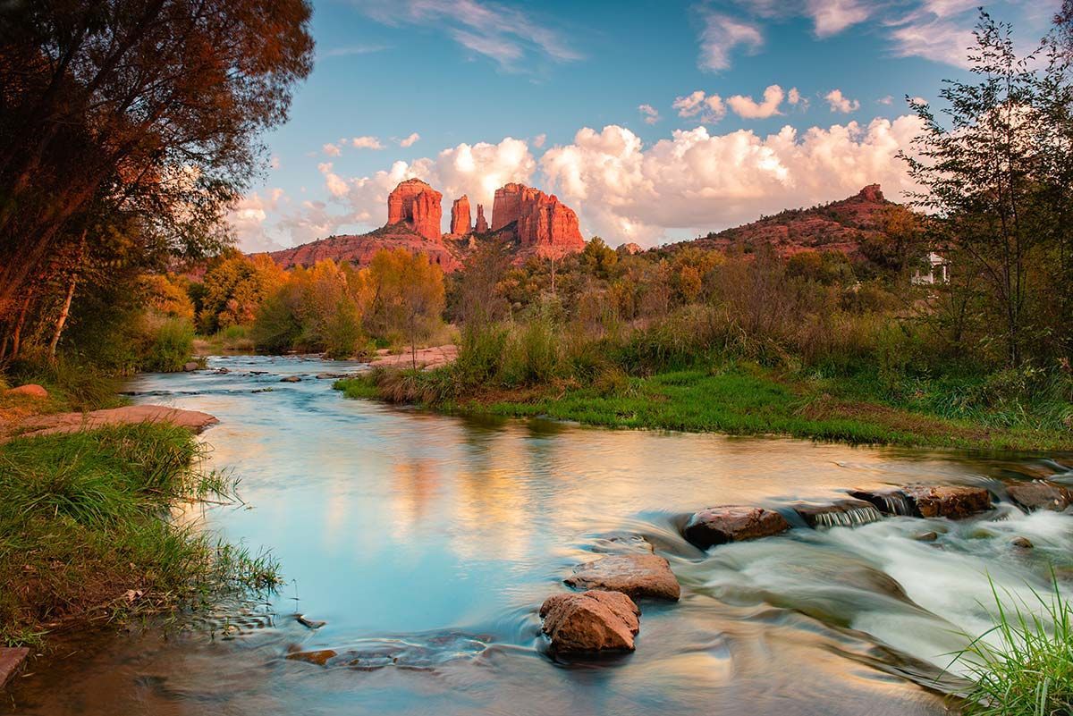 River flows through a scenic landscape; red rock formations and colorful sky in the background.