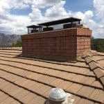 Brick chimney on a tiled roof with protective screens, set against a backdrop of a mountain landscape.