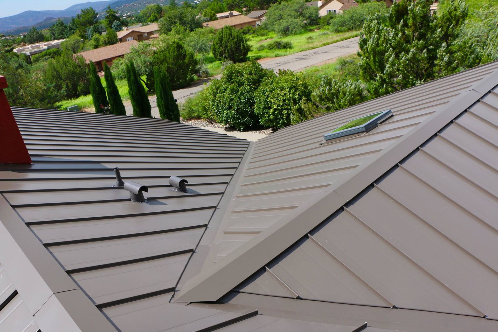 Metal roof, brown panels, chimney, pipes, green foliage and houses in background.