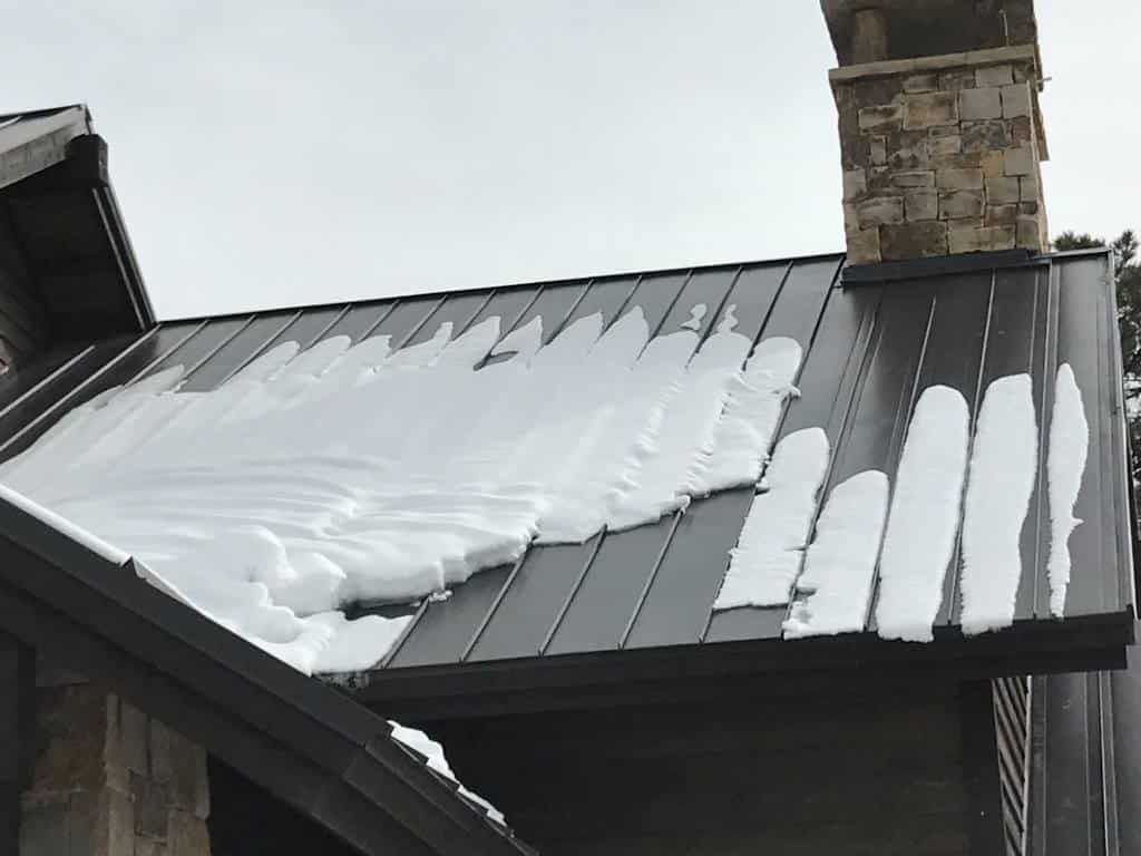 Snow-covered dark metal roof with strips of melting snow, next to a stone chimney.