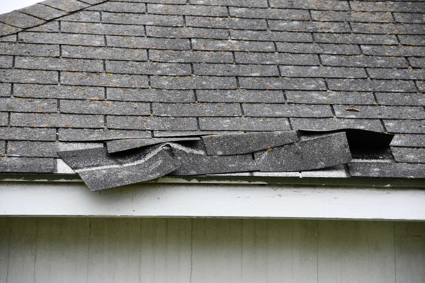 Damaged asphalt shingle roof, with missing and curled shingles along the edge of a white building.