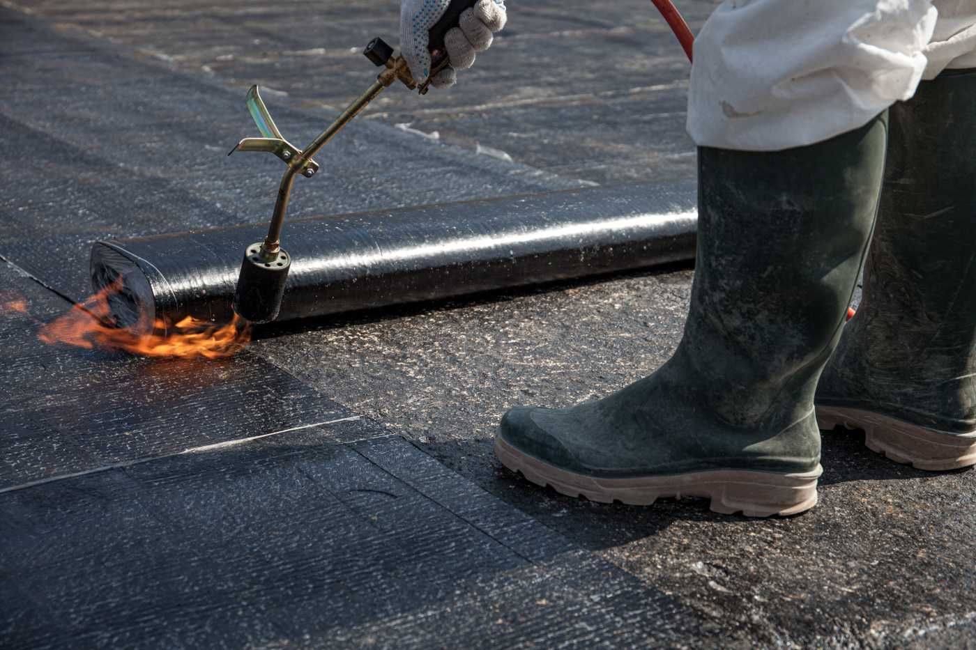 Person in boots using a torch to seal black roofing material outdoors.