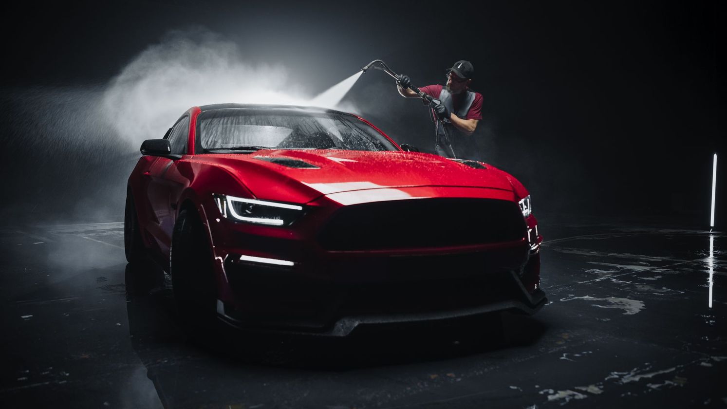 A red sports car being washed with a pressure washer, person in background, dark setting.