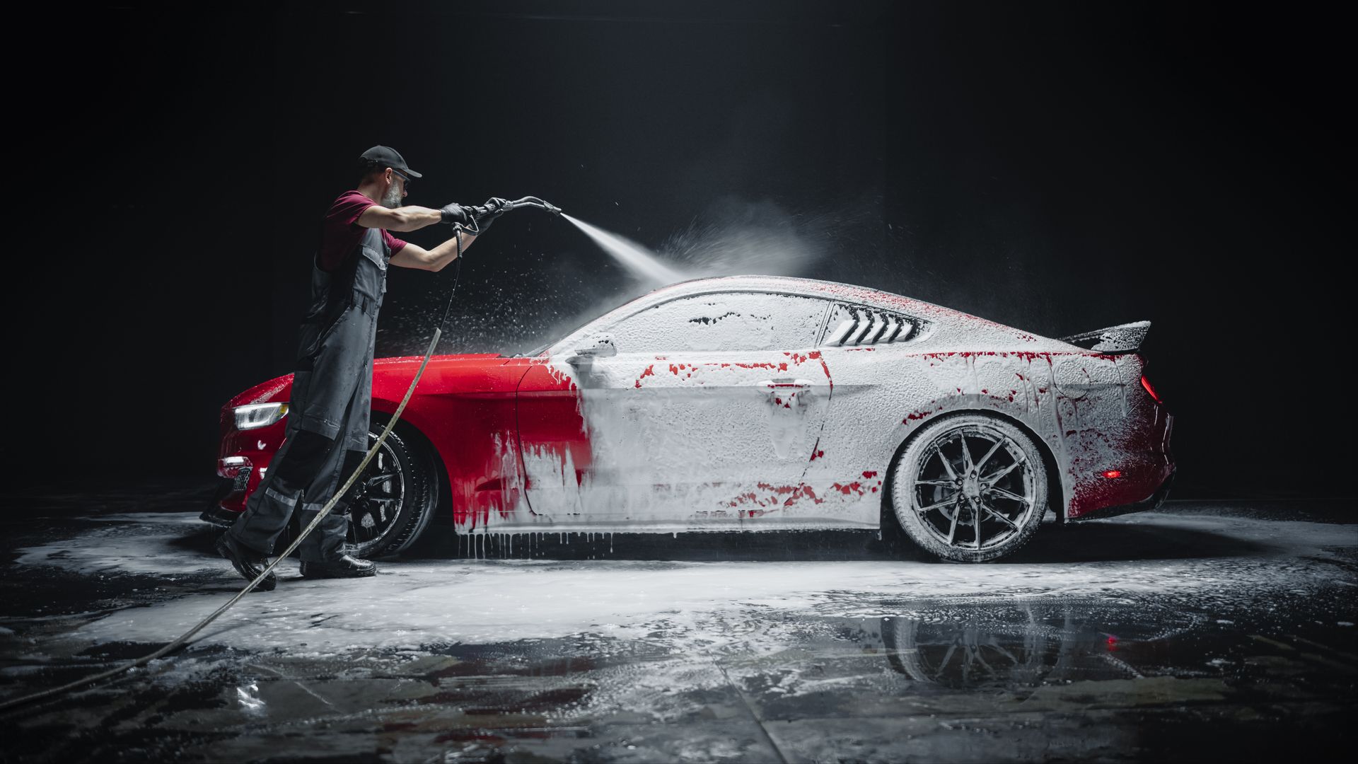 Person washing a red and white sports car with a pressure washer in a dark setting.