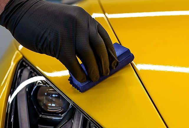A gloved hand uses a blue applicator pad to apply ceramic coating to a bright yellow car hood.