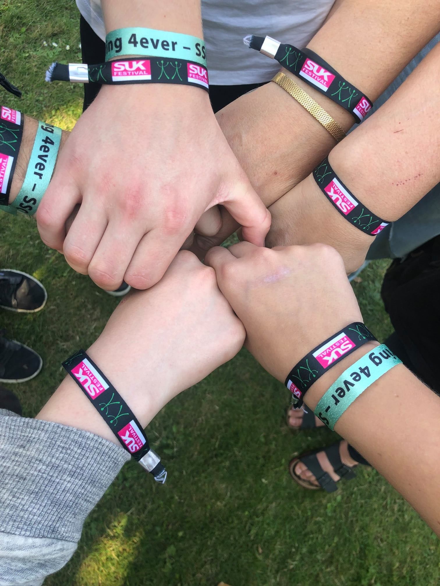 Hænder i en cirkel med matchende sorte og blågrønne armbånd, muligvis ved et arrangement på græs.