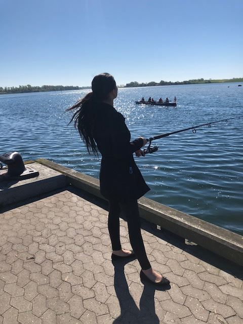 Person, der fisker på en kaj, ser et rohold på vandet; solskinsdag.