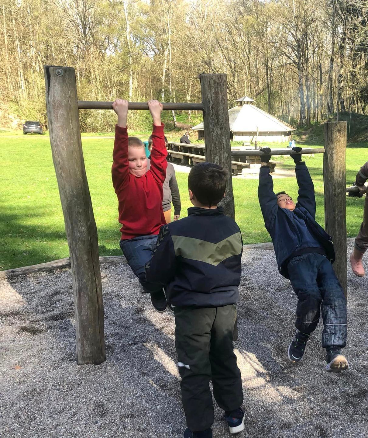 Tre børn leger på klatrestativer i en park. En i rød skjorte hænger. De andre gynger.