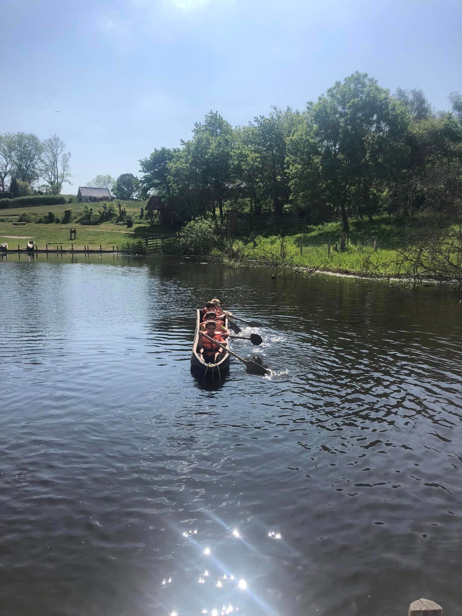 Folk padler i kano på en sø med træer og grønt græs på bredden. Solskinsdag.