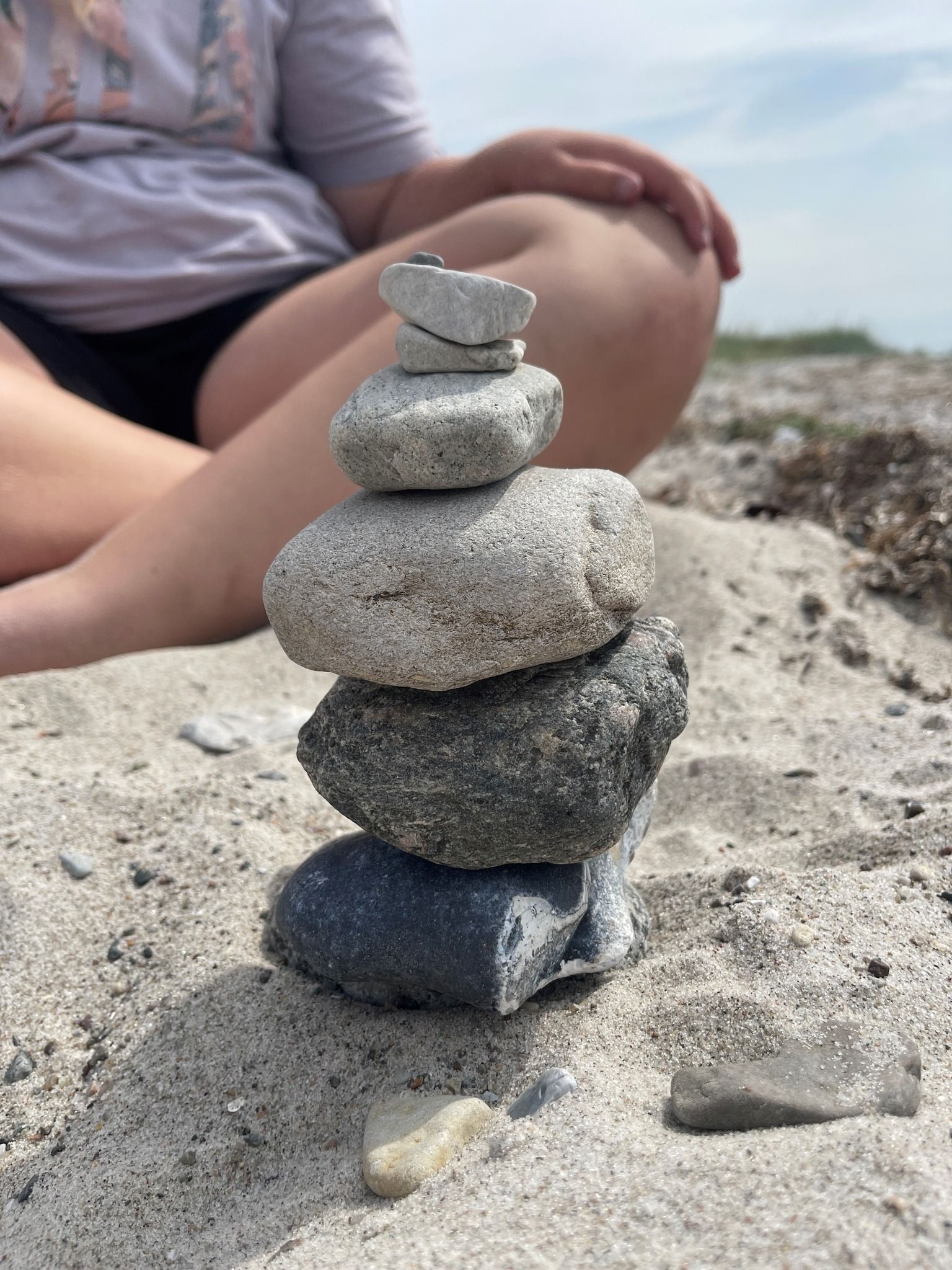 Stak af grå sten balanceret på sand, med en persons ben i baggrunden.
