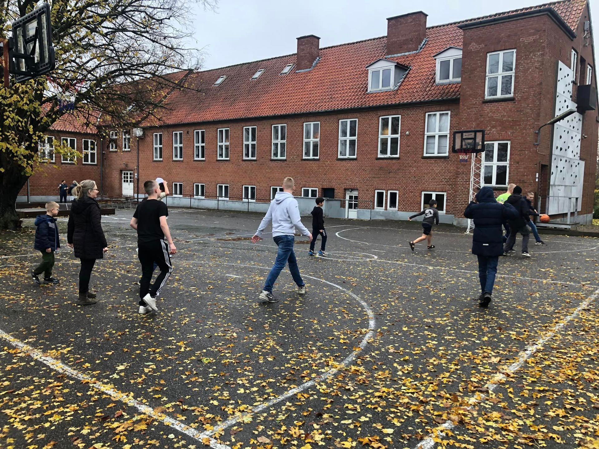 Børn spiller basketball på en betonbane dækket af efterårsblade foran en murstensskolebygning.