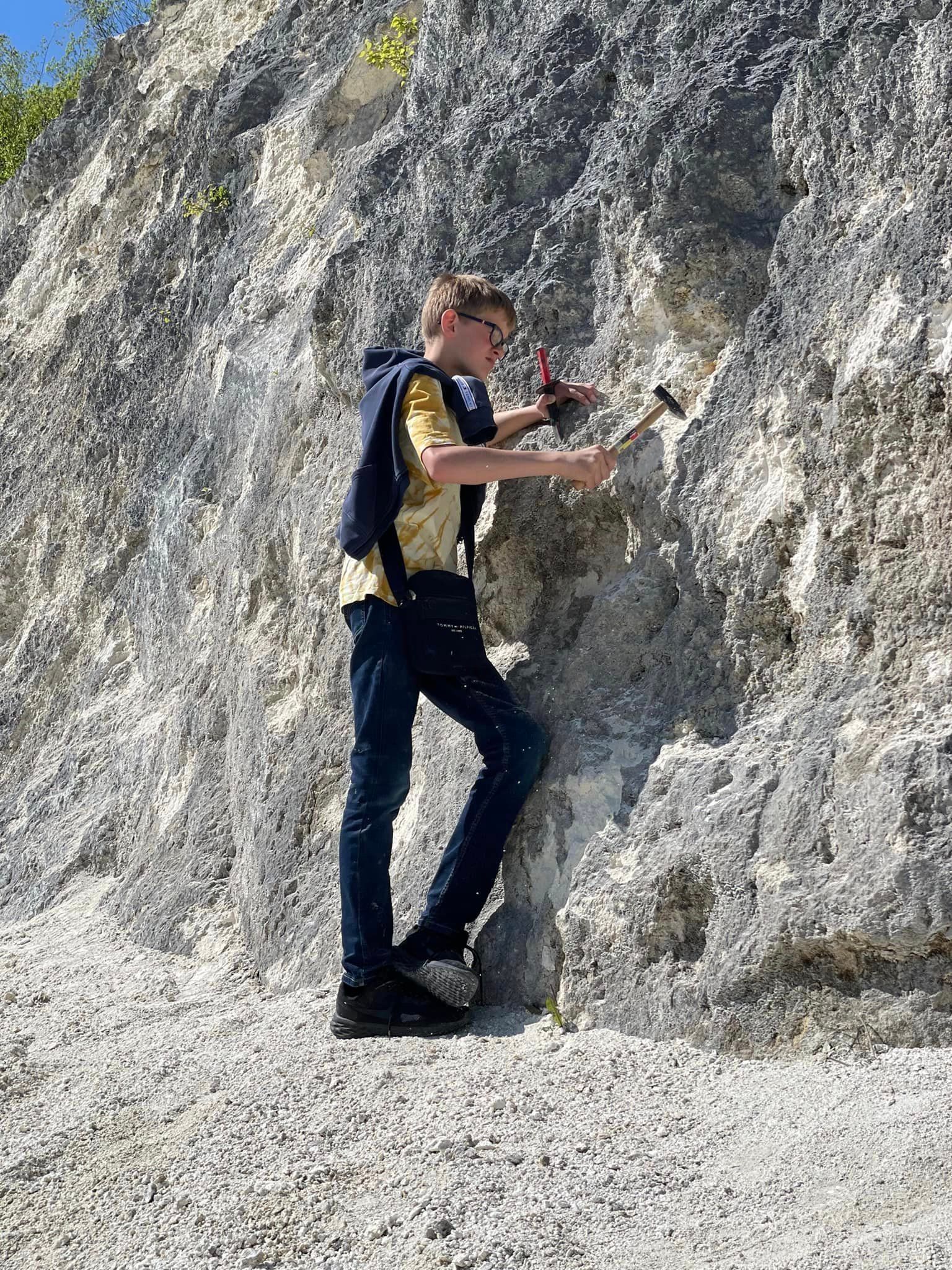 Dreng bruger hammer på klippevæg i et lyst stenbrud. Han har briller, jakke og jeans på.