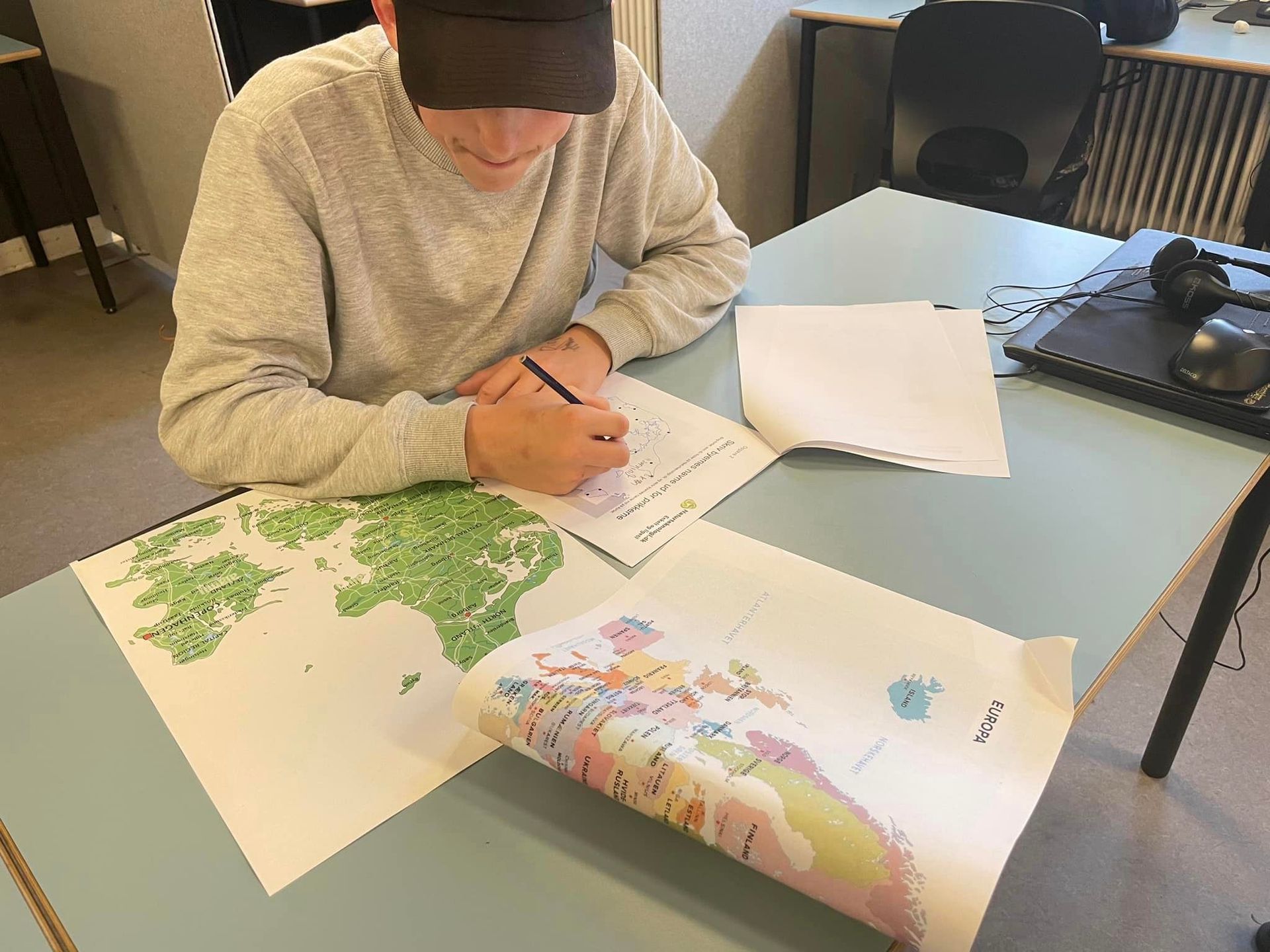Person skriver ved et skrivebord med kort og papirer. Iført kasket og lys sweater.